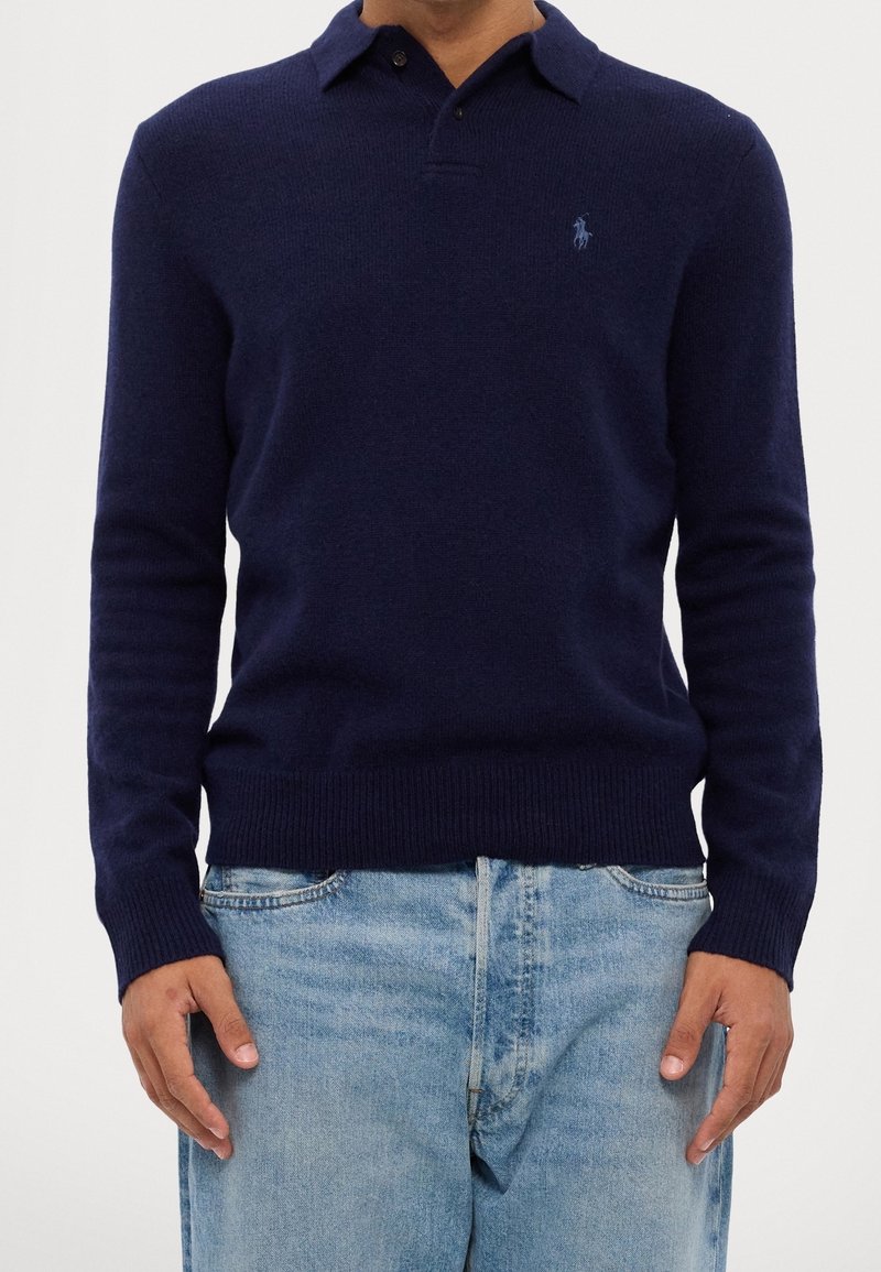 Homme portant un pull polo bleu marine à manches longues et un jean bleu clair, les mains détendues sur les côtés.