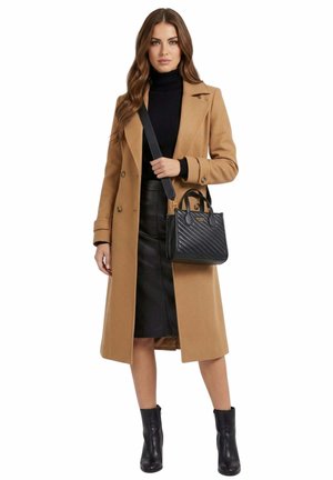 Femme portant un manteau camel, un col roulé noir, une jupe noire, des bottines, tenant un sac à main noir Guess avec bandoulière.