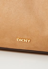 Hnědá semišová kabelka s hladkým povrchem, která na přední straně nese výrazné zlaté logo "DKNY". Klasický design s čistými liniemi.