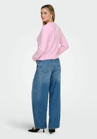 Pull rose à manches longues associé à un jean bleu large à taille haute. La tenue présente un design simple et une texture douce. Des talons noirs complètent le look.