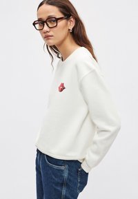 Felpa bianca con grafica di labbra rosse sul petto, con vestibilità rilassata e scollo rotondo, abbinata a jeans di denim blu.