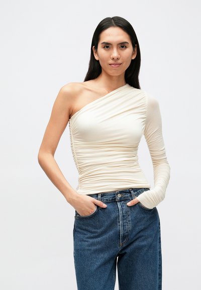 Top creme de um ombro só com textura franzida, manga comprida justa, combinado com jeans de cintura alta azul escuro, criando um visual contrastante.