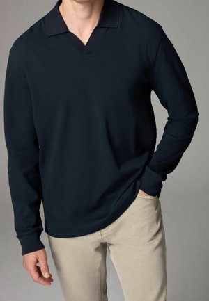 Homme portant un polo noir à manches longues avec un pantalon beige, une main dans la poche et l'autre détendue le long du corps.