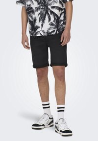Pantalones cortos de mezclilla negra con dobladillos enrollados, combinados con una camisa de estampado de hojas de palmera en blanco y negro, calcetines blancos con rayas negras y zapatillas.
