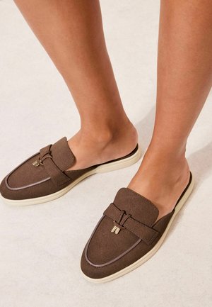 Stopy w brązowych wsuwanych mokasynach z kremowymi podeszwami, zdobione ozdobnym paskiem i złotymi akcentami, stojące na jasnej podłodze.