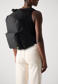 Sac à dos en tissu noir au design épuré, doté d'une poche avant et d'un détail logo jaune. Associé à un jean clair.