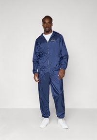 Marineblauwe windbreaker en bijpassende broek gemaakt van lichtgewicht, glanzend materiaal. Heeft een rits aan de voorkant en elastische mouwen; gedragen met witte sneakers.