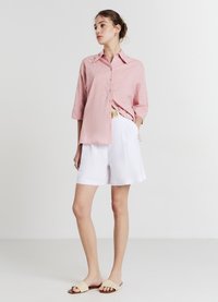 Camicia rosa chiaro con bottoni e motivo testurizzato, maniche a tre quarti; abbinata a pantaloni shorts bianchi fino al ginocchio e sandali beige a scivolo.
