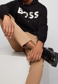 Uomo che indossa una felpa nera "BOSS", pantaloni beige e scarpe di pelle nere, seduto con una gamba incrociata sopra l'altra su un blocco bianco.