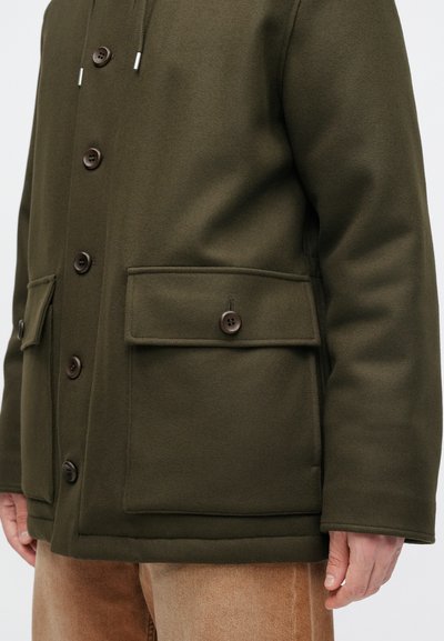 Oliivinvihreä villasekoitepäällystakki, jossa on suuret etutaskut, napit sulkemiseen ja pehmeä tekstuuri, rento istuvuus ja minimalistinen muotoilu.