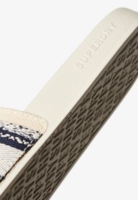 Slide-sandalen met een gestructureerde witte zool. De bovenkant heeft een geweven patroon in marineblauw en beige, naast een merkdetail op de zool.