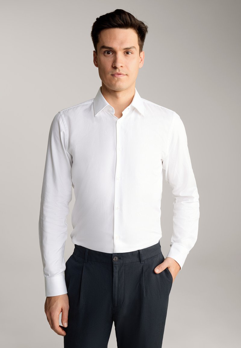 Camicia bianca con bottoni e maniche lunghe, ben aderente, abbinata a pantaloni scuri. Tessuto liscio, colletto classico e polsini con bottoni.