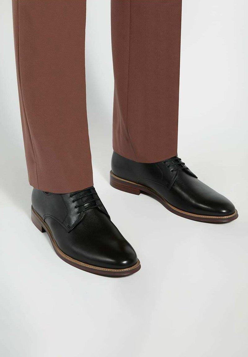 Des chaussures de ville en cuir noir au fini lisse, avec un design à lacets et une semelle marron, associées à un pantalon marron ajusté.