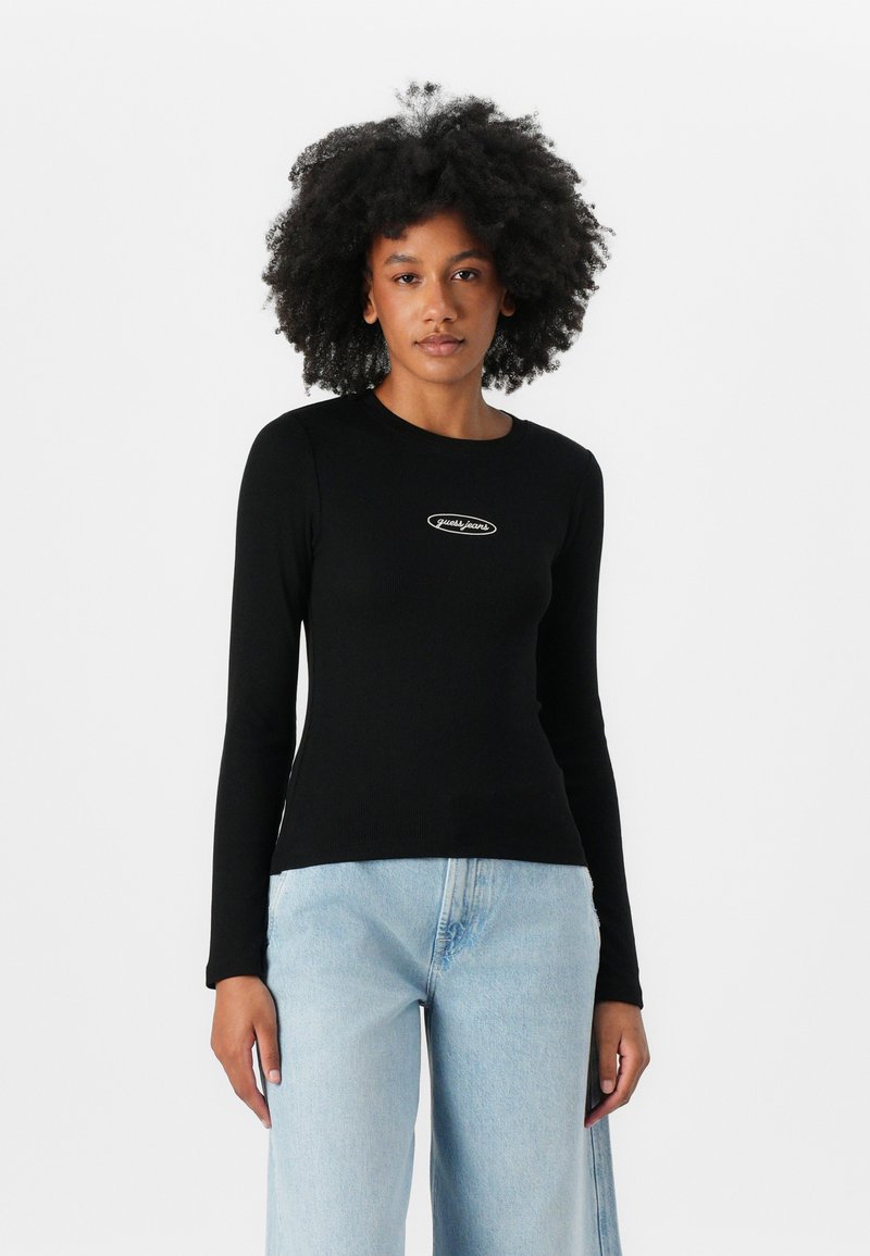 Svart, langermet tettsittende topp med rund hals og ribbestruktur, med en liten hvit logo på brystet. Kombinert med lyseblå jeans.