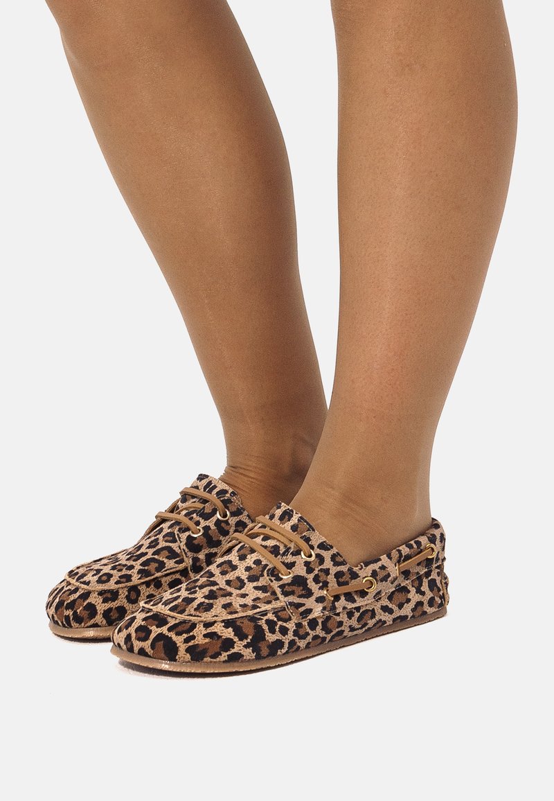Pies con mocasines de estampado de leopardo tipo slip-on, con cordones color canela y ojales dorados, sobre fondo blanco.