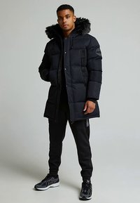 Άνδρας που στέκεται φορώντας ένα μαύρο χοντρό χειμερινό παλτό με γουναριστή κουκούλα, μαύρα παντελόνια φόρμας και μαύρα αθλητικά παπούτσια σε ελαφρύ γκρι φόντο.