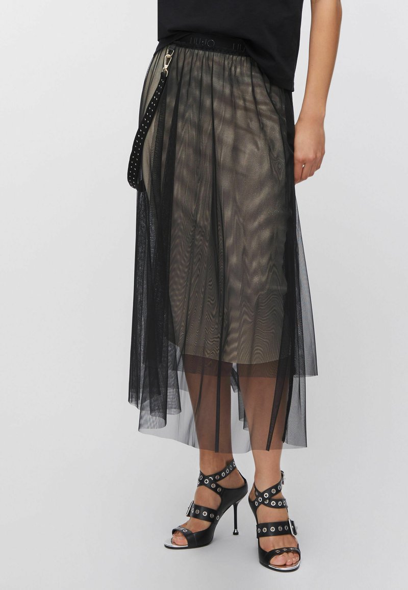 Gonna in tulle nero trasparente con design fluido e vita regolabile. Abbinata a sandali neri con cinturino e dettagli metallici.