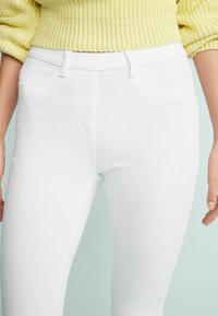 Białe dżinsy skinny z przodu kieszeniami i szlufkami na pasek, o gładkiej fakturze i starannym wykończeniu. W komplecie z żółtym swetrem dzianinowym.