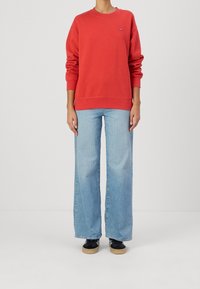 Sudadera roja con puños y dobladillo acanalados, combinada con vaqueros de pierna ancha de color azul claro. Zapatos negros con suelas marrones completan el conjunto.