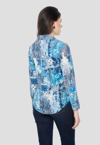 Modrá košeľa s dlhými rukávmi na gombíky s komplikovanými vzormi a textúrami. Má špicatý golier a zaoblený spodný okraj.