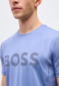 Κοντινό πλάνο ενός ατόμου που φορά ένα ανοιχτό μπλε T-shirt με την επιγραφή "BOSS" σε μεγάλα ριγωτά γράμματα στο στήθος, με ασημένιο κρίκο σκουλαρίκι ορατό.