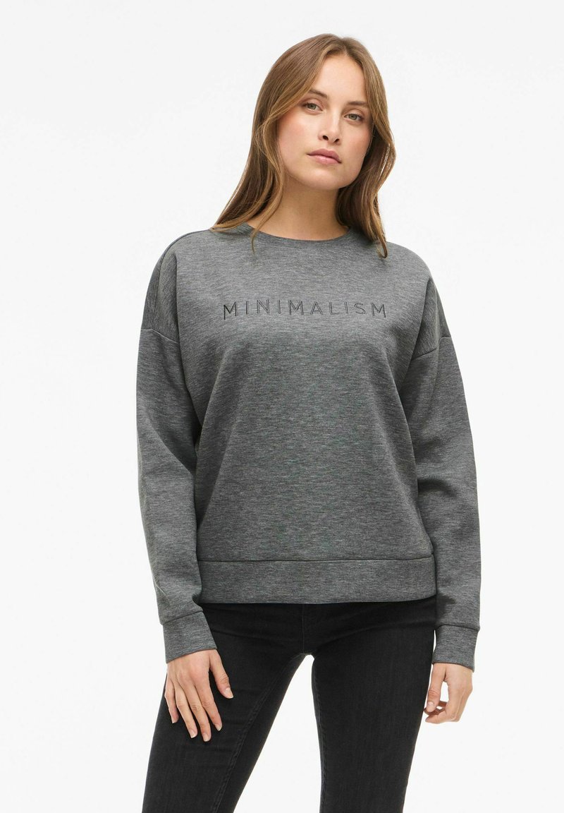 Žena se světle hnědými vlasy, oblečená v šedé mikině s nápisem "MINIMALISM" a černými kalhotami, stojí na bílém pozadí.