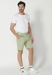 Pantalones cortos verde claro con cinturilla elástica, bolsillos laterales y textura suave; combinados con una camiseta blanca simple y zapatillas blancas.