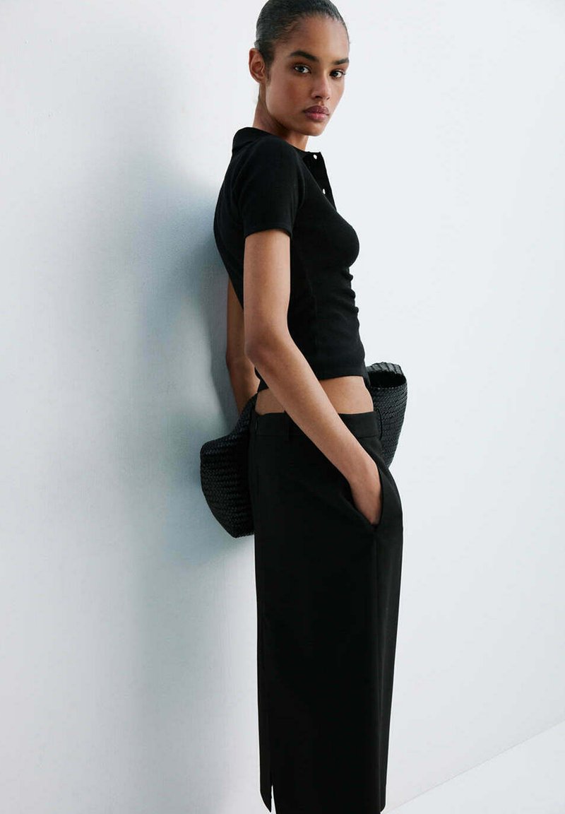 Femme portant un haut noir ajusté à manches courtes et un pantalon noir taille haute, debout de profil avec une main dans la poche et un sac noir derrière elle.