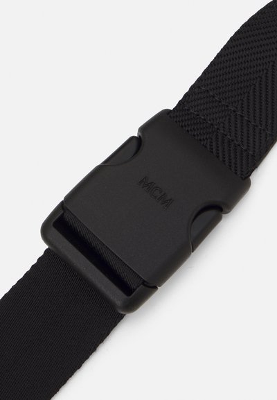 Cinghia intrecciata nera con una fibbia a rilascio laterale in plastica nera, incisa con il logo del marchio "MCM" su uno sfondo bianco.