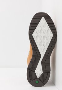 Suola di scarpa in gomma marrone con un motivo di battistrada bianco a forma di croce, superficie testurizzata e logo del marchio visibile, appoggiata su uno sfondo bianco.