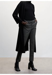 Sort lang cardigan over en sort ribbet rullekrave, parret med grå ternede bukser og sorte hælede ankelstøvler. Glatte teksturer, skræddersyet pasform.