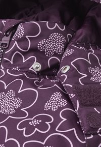 Lilla jakke stof med stort blomstermønster i lys pink og hvid, har en lynlås, trykknap og velcro detaljer.