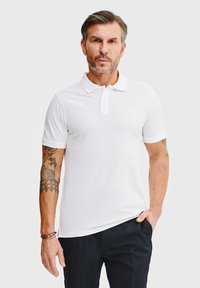 Biała koszulka polo z klasycznym kołnierzykiem, z trzema guzikami, krótkimi rękawami i gładką teksturą, w połączeniu z ciemnymi spodniami.