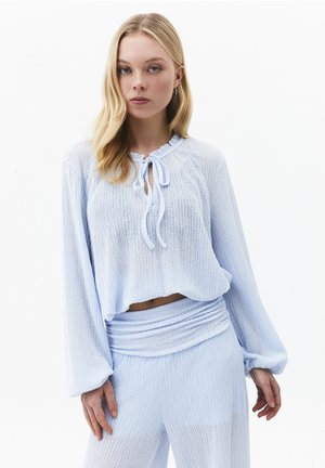 Blonde vrouw die een lichtblauw, gestructureerd topje met lange mouwen en strikkoordje bij de hals draagt, gecombineerd met bijpassende high-waisted broek, staand tegen een witte achtergrond.