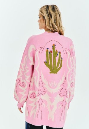 Kobieta o blond włosach, ubrana w różowy sweter z dużym zielonym kaktusem i małymi kolorowymi kwiatami na plecach.