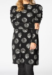 Robe noire à manches longues, avec des motifs circulaires blancs en forme de feuilles. Épaules bouffantes et encolure volantée ajoutent de la texture et des détails.