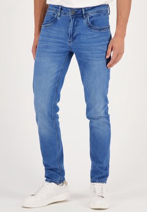 Schmale blaue Denim-Jeans, getragen mit weißen Turnschuhen, sichtbar von der Taille bis zu den Füßen vor einem schlichten Hintergrund.