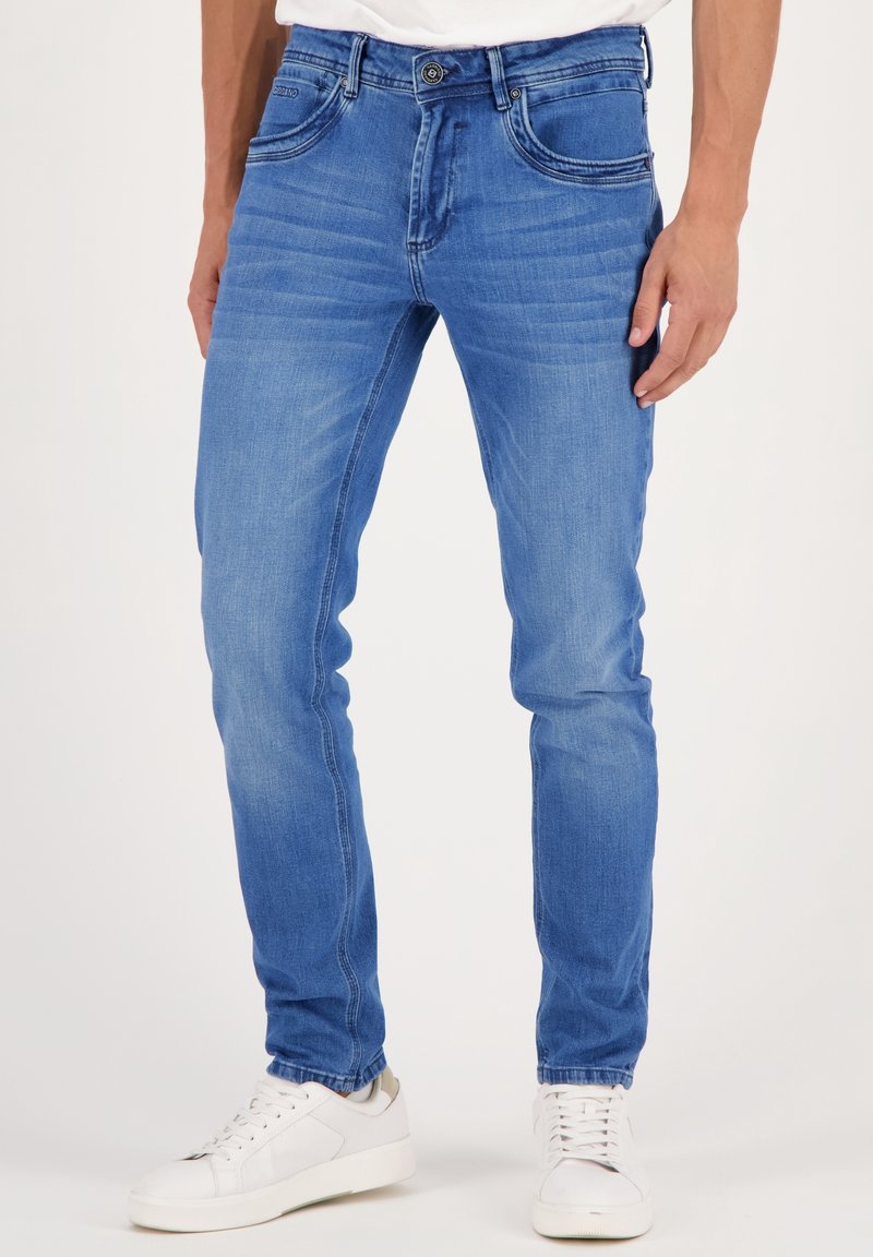 Jean en denim bleu coupe slim porté avec des baskets blanches, visible de la taille aux pieds sur un fond uni.
