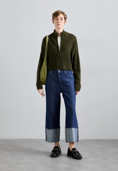 Woman wearing an olive green zip-up sweater, wide cuffed blue jeans, black loafers, and carrying a green shoulder bag, standing against a plain wall.