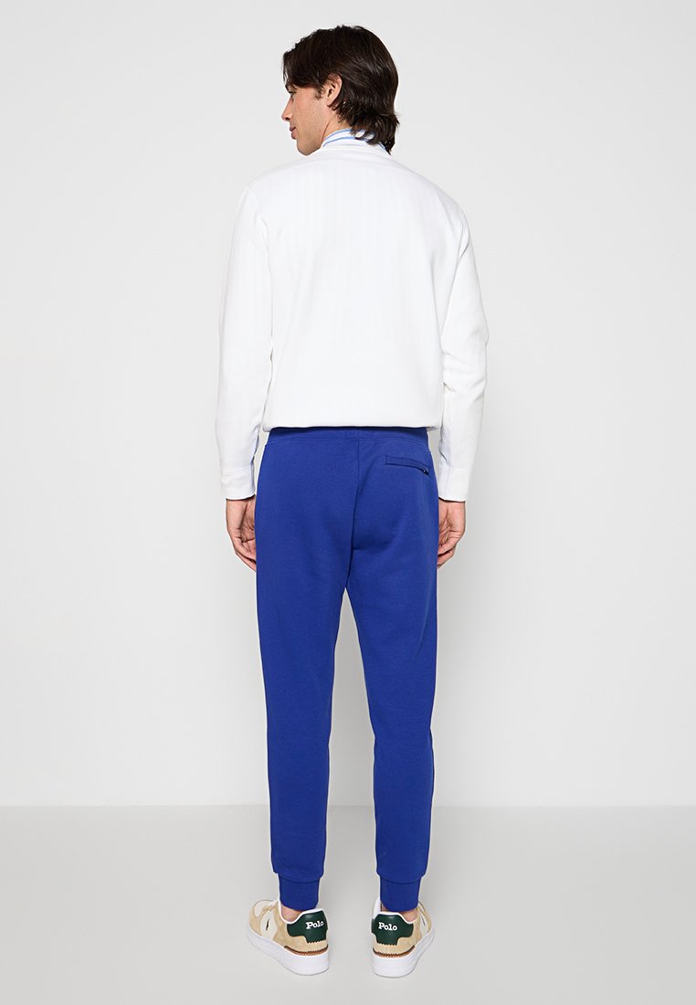 Niebieskie dresy o dopasowanym kroju z detalami na tylnej kieszeni, w połączeniu z białą koszulą z długimi rękawami. Widoczne swobodne obuwie.
