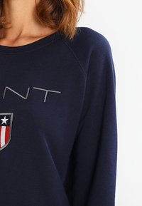 Granatowa bluza z okrągłym dekoltem, z szarym napisem i herbem z czerwonymi, białymi i niebieskimi akcentami. Miękki, teksturowany materiał.