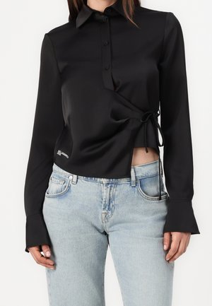 Frau trägt eine schwarze langärmelige Bluse mit Knöpfen und seitlichem Bindeband sowie hellblaue Jeans, steht vor einem schlichten Hintergrund.
