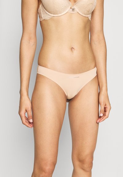 Fio dental bege claro feito de material suave, com cobertura mínima e um detalhe com o logótipo na cintura. Textura suave, design sem costuras.