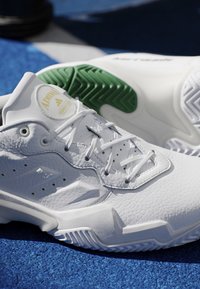 Scarpe da tennis Adidas bianche con suole verdi e bianche appoggiate su una superficie blu, che mostrano texture e lacci dettagliati.