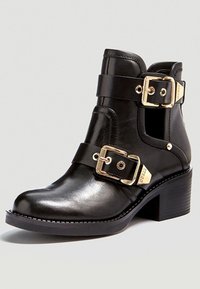 Bottine en cuir noir avec des boucles dorées, bout arrondi et talon épais. Présente une texture lisse et des accents de sangle décoratifs.