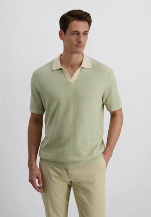 Mann mit braunen Haaren, der ein hellgrünes kurzärmeliges Poloshirt aus Strick und beige Hose trägt, steht mit einer Hand in der Tasche.