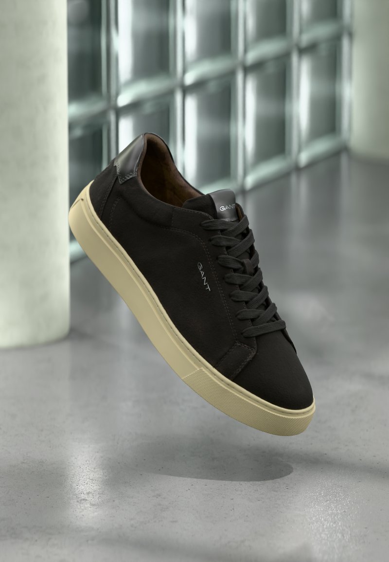 Schwarze Wildleder-Sneaker mit niedrigem Profil, heller Gummisohle, kontrastierenden schwarzen Lederakzenten an der Ferse und schwarzen Schnürsenkeln.
