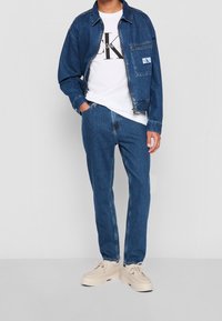 Jeansjacka med två framfickor, bärs över en vit T-shirt med en stor svart logo. Blå raka jeans och ljusa sneakers.