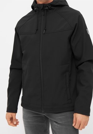 Mężczyzna ubrany w czarną kurtkę z kapturem i suwakiem, z przednimi kieszeniami i regulowanymi troczkami, noszący szare dżinsy.