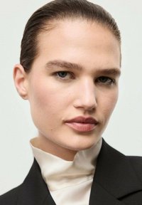 Primo piano di una persona con pelle liscia, capelli castano chiaro, trucco neutro, che indossa un dolcevita beige sotto un giubbotto nero. Sfondo chiaro.